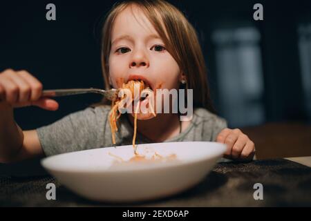 Hungrige kleine Mädchen Appetit essen die Bolognese Pasta aufrollend es auf der Gabel und setzen auf den Mund in der niedrigen Licht Küchentisch. Stockfoto