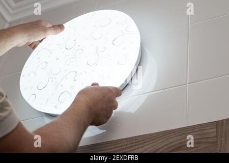 Ein Mann installiert eine große runde weiße LED-Lampe an der Wand im Badezimmer. Inneneinrichtung und Wohnungsbau. Stockfoto