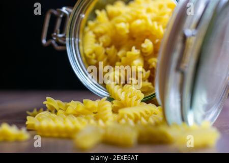 Fussili Pasta aus einem großen luftdichten Glas fallen. Rohe trockene Nudeln in Null-Abfall-Glasbehälter. Chaotisch Lebensmittelkomposition auf dunklem Hintergrund mit Stockfoto