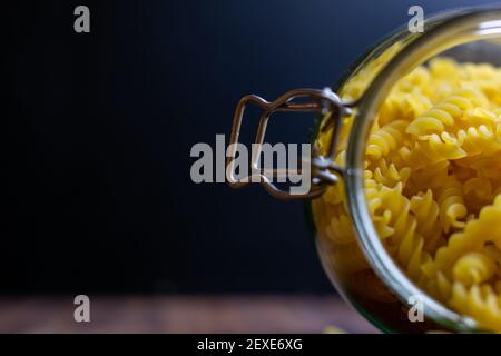 Fussili Pasta aus einem großen luftdichten Glas fallen. Rohe trockene Nudeln in Null-Abfall-Glasbehälter. Chaotisch Lebensmittelkomposition auf dunklem Hintergrund mit Stockfoto