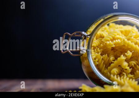 Fussili Pasta aus einem großen luftdichten Glas fallen. Rohe trockene Nudeln in Null-Abfall-Glasbehälter. Chaotisch Lebensmittelkomposition auf dunklem Hintergrund mit Stockfoto