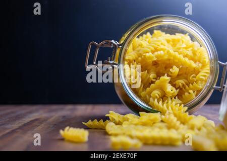 Fussili Pasta aus einem großen luftdichten Glas fallen. Rohe trockene Nudeln in Null-Abfall-Glasbehälter. Chaotisch Lebensmittelkomposition auf dunklem Hintergrund mit Stockfoto