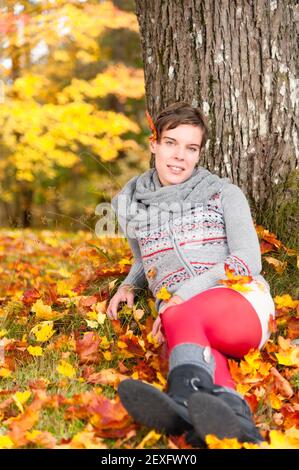 Die junge Frau sitzt im Herbst an einem Baum gelehnt Stockfoto