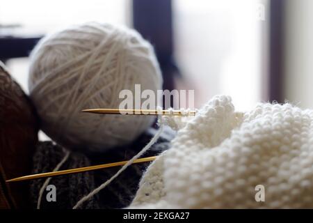 Weiße Schafwolle, die zu einem Ball gewickelt ist, neben gelben Stricknadeln und Strickstoff. Nahaufnahme, selektiver Fokus. Handarbeitskonzept, altmodisch Stockfoto