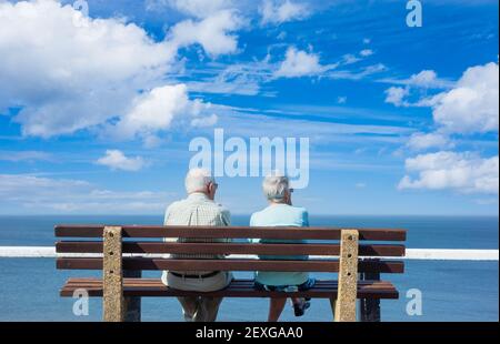 Rückansicht eines älteren Ehepaares, das an einem sonnigen Tag auf dem Sitz mit Blick auf das Meer sitzt. Stockfoto