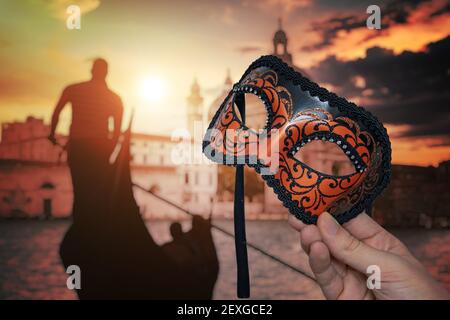 Hand hält venezianische Karnevalsmaske auf traditionellen venezianischen Festival in Italien. Gondel im Hintergrund bei Sonnenuntergang. Stockfoto