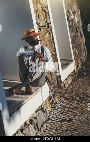 Ein Porträt eines modischen schwarzen Kerl in einem Hut und ein elegantes Outfit sitzen auf einer Schwelle im Freien und gehen, um eine Zigarre nach einem guten Golfspiel zu rauchen, zwei Stockfoto