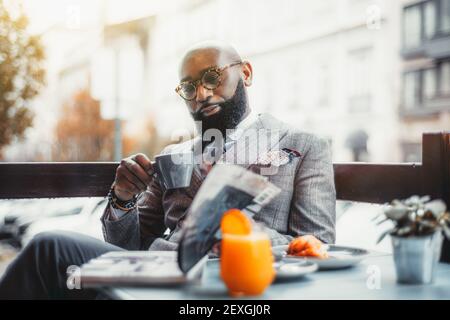 Porträt eines baldheaded erwachsenen schwarzen Dandy Kerl mit einem gepflegten Bart, in einem maßgefertigten eleganten Kostüm und Brillen sitzen in einem Straßencafe Witz Stockfoto