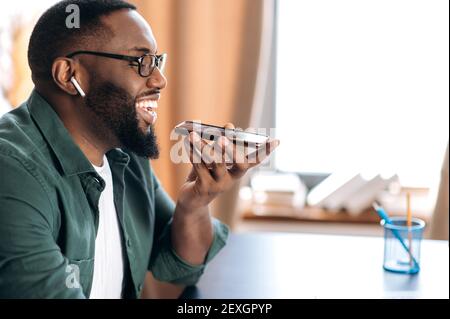 Nahaufnahme eines attraktiven zufriedenen afroamerikanischen Kerl, halten und mit Handy, sprechen mit Freund oder Kollegen Freisprecheinrichtung, sitzen zu Hause oder im Büro Stockfoto