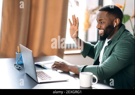 Online-Kommunikation. Glücklich lächelnder afroamerikanischer Kerl, Freiberufler oder Student, winkende Hand, Gruß, verwendet Laptop für Videokonferenz mit Kollegen Stockfoto