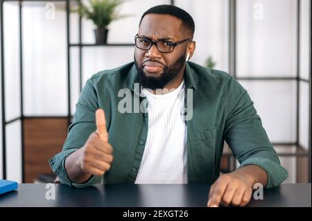 Porträt eines freundlichen, erfolgreichen bärtigen Afroamerikaners mit Brille, der an einem Tisch sitzt und mit Daumen nach oben zeigt und direkt in die Kamera blickt Stockfoto