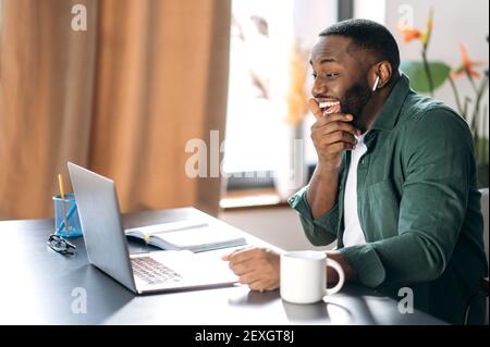 Fröhlicher afroamerikanischer Kerl in stilvoller Kleidung verwendet einen Laptop für Online-Arbeit, sitzt an seinem Schreibtisch, schaut glücklich auf den Bildschirm, bekam eine gute Nachricht Stockfoto
