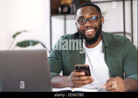 Porträt eines freundlichen afroamerikanischen jungen Mannes, der eine Brille trägt, am Schreibtisch sitzt, Smartphone hält und mit einem Lächeln direkt in die Kamera blickt Stockfoto