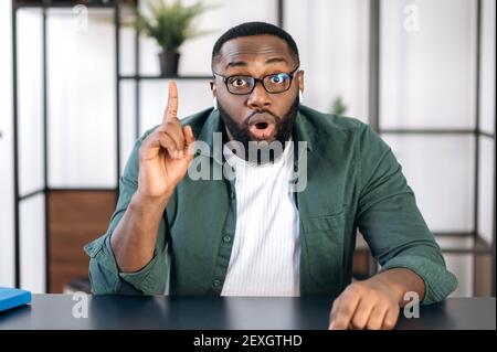 Porträt eines aufgeregt schockierten bärtigen afroamerikanischen Typen mit Brillen, der an einem Tisch sitzt und eine Idee zeigt und direkt in die Kamera schaut Stockfoto
