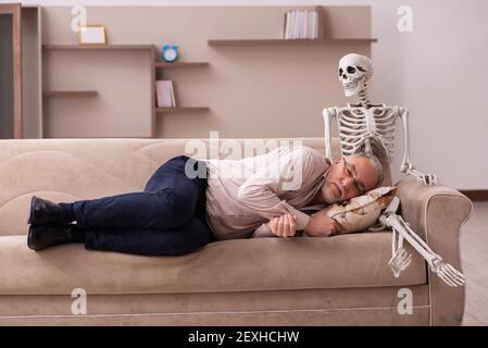 Alter Mann sitzt auf dem Sofa mit dem weiblichen Skelett In unsterbst Liebe Konzept Stockfoto