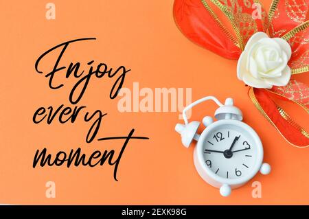 Uhr und Blume auf orangefarbenem Hintergrund mit JEDEM MOMENT GENIESSEN geschrieben. Stockfoto