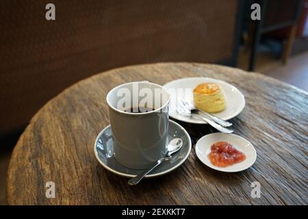 Eine Tasse heißen schwarzen Kaffees mit Butter serviert Schlichte Scone und Beerenmarmelade auf Holztisch Stockfoto