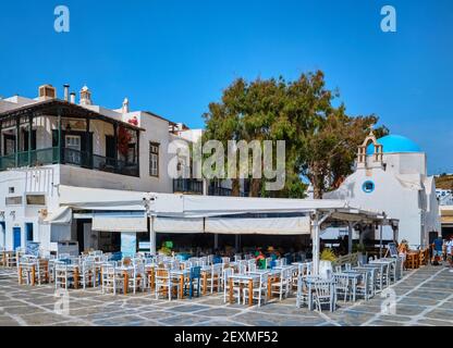 Farbenprächtiges Café oder Restaurant im Freien in einer typisch griechischen Inselstadt. Griechisch-orthodoxe Kirche, blaue Kuppel, Touristen herumlaufen. Mykonos, Kykladen, Griechenland. Stockfoto