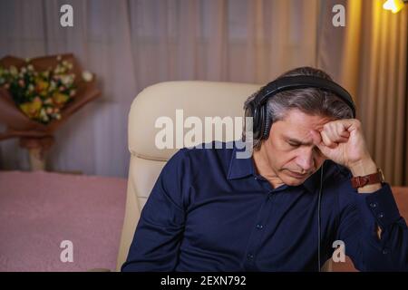 Der 50-jährige Mann hört zu Hause Musik auf Kopfhörern, sitzt auf einem Stuhl. Erholsame Freude. Stockfoto