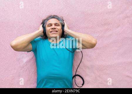 Der 50-jährige Mann hört Musik mit Kopfhörern zu Hause, auf dem Bett liegend. Entspannend, genießend. Stockfoto