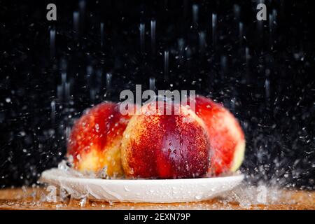 Stillleben von drei reifen Pfirsichen in einem Teller auf einem hölzernen Tisch unter Wasserspritzern auf einem schwarzen isolierten Hintergrund. Hochwertige Fotos Stockfoto
