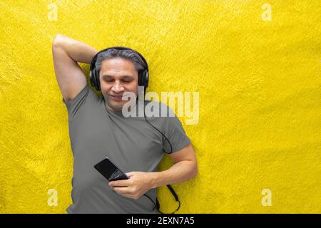 Der 50-jährige Mann hört Musik mit Kopfhörern zu Hause, auf dem Bett liegend. Entspannend, genießend. Stockfoto