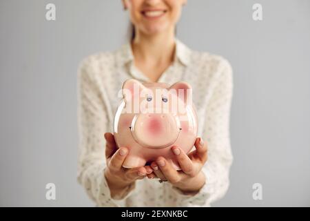 Nahaufnahme eines Sparschweins in Form eines rosa Schweins in den Händen einer Frau auf grauem Hintergrund. Stockfoto