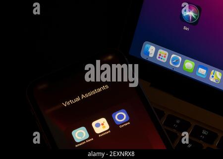 Virtual Assistant-Apps werden auf einem iPhone und einem MacBook Air angezeigt - Amazon Alexa, Google Assistant, Cortana und Siri. Stockfoto