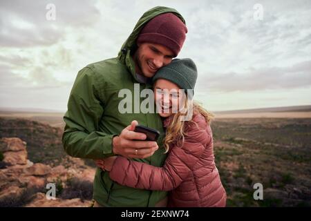 Lachend junges Paar umarmt während des Lagers auf den Bergen Blick auf Smartphone in Winterkleidung Stockfoto