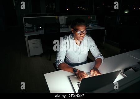 Afrikanischer junger Geschäftsmann, der bis spät in die Nacht am Laptop arbeitet Nachts im dunklen Büro Stockfoto