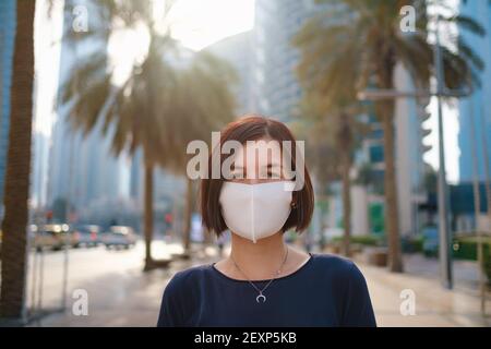 Portrait der jungen Frau trägt in blauem Kleid und weiße Maske für Virus zu verhindern, gehen Sie vor Skycrapers in der modernen Stadt. Stockfoto