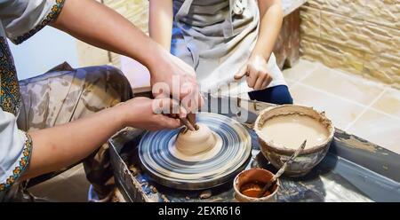 Die Hände eines Meisters und eines Studenten machen einen Krug auf einem Potter-Rad aus gelbem Ton. Selektive Fokussierung auf die Hände Stockfoto