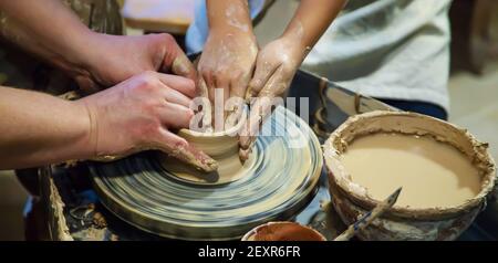 Die Hände eines Meisters und eines Studenten machen einen Krug auf einem Potter-Rad aus gelbem Ton. Selektive Fokussierung auf die Hände Stockfoto