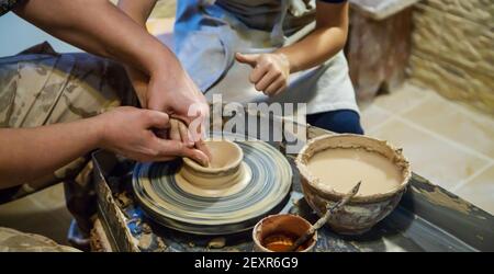 Die Hände eines Meisters und eines Studenten machen einen Krug auf einem Potter-Rad aus gelbem Ton. Selektive Fokussierung auf die Hände Stockfoto
