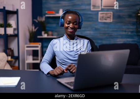 Schwarze Frau in guter Laune tragen Headset Musik hören arbeiten fristgerecht aus dem Home Office. Sitzen am Schreibtisch. Afrikanischer Freiberufler, der neues Projekt erstellt, das spät arbeitet. Stockfoto