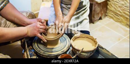 Die Hände eines Meisters und eines Studenten machen einen Krug auf einem Potter-Rad aus gelbem Ton. Selektive Fokussierung auf die Hände Stockfoto