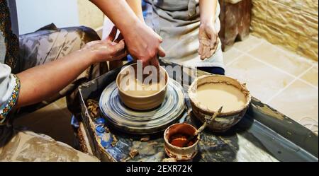 Die Hände eines Meisters und eines Studenten machen einen Krug auf einem Potter-Rad aus gelbem Ton. Selektive Fokussierung auf die Hände Stockfoto
