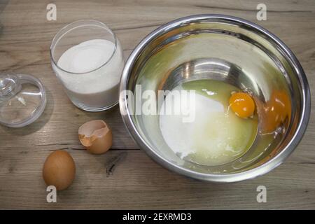 Weißer Zucker und Ei in einer Schüssel und zwei Eier auf dem Holzhintergrund. Der zweite Schritt für die Herstellung von Muffins, Kuchen, Keks. Backvorgang Schritt für Schritt. Stockfoto