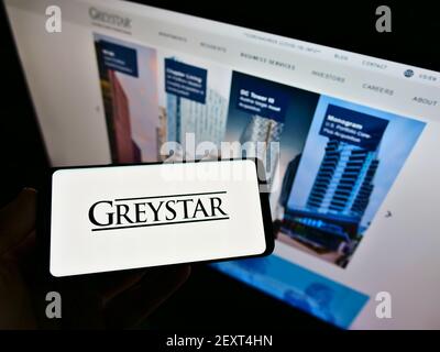 Person hält Handy mit Logo der US-Wohnungsgesellschaft Greystar Real Estate Partners auf dem Bildschirm vor der Webseite. Fokus auf Handy-Display. Stockfoto
