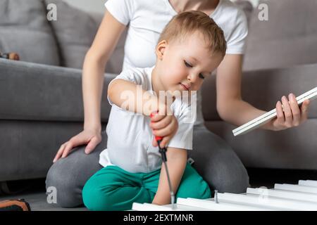 Cute liebenswert konzentriert kaukasischen Kleinkind Junge Kind sitzen auf dem Boden und helfen Mama Montage Möbel Regal mit Schraubendreher. Junge Erwachsene Mutter mit lustig Stockfoto