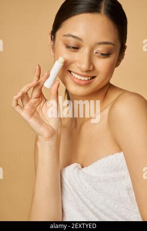 Attraktive junge Frau in Handtuch eingehüllt lächelnd und hält Lippenbalsam während posiert isoliert über beige Hintergrund. Konzept für Schönheitsbehandlungen Stockfoto