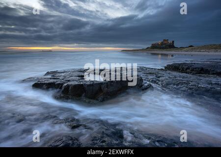 Bamburgh Castle überblickt die Nordseeküste von Northumberland unter dunklen und brütenden Wolken bei Sonnenaufgang an einem frühen Frühlingsmorgen. Stockfoto