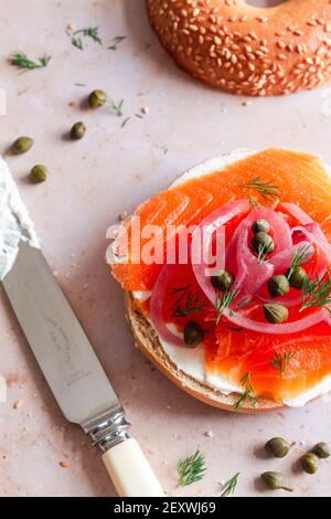 Bagel zur Hälfte mit Frischkäse und gekrönt mit geräucherter Forelle, eingelegten roten Zwiebeln, Kapern und Dill. Stockfoto
