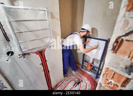 Klempner Installation verdeckter Toilettenrahmen im Badezimmer. Junger Mann im Schutzhelm, der die Abdeckung auf dem Rahmen platziert, während er ein an der Wand hängende Toilettensystem installiert. Konzept der Sanitär-Arbeiten und Renovierung. Stockfoto