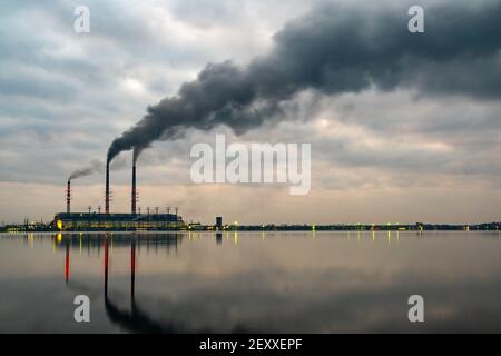 Kohlekraftwerk hohe Rohre mit schwarzem Rauch, der nach oben die verschmutzende Atmosphäre mit den Reflexionen von ihm im Seewasser bewegt. Stockfoto