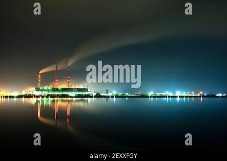 Hell erleuchtetes Kohlekraftwerk hohe Rohre mit schwarzem Rauch, der nach oben die verschmutzende Atmosphäre in der Nacht mit den Reflexen von ihm im Seewasser bewegt. Stockfoto