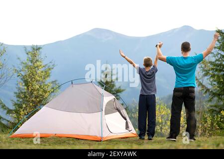 Der junge Vater und sein kleiner Sohn stehen in der Nähe des Campingzeltes mit erhobenen Händen beim gemeinsamen Wandern in den Sommerbergen. Konzept für aktives Familienreisen. Stockfoto