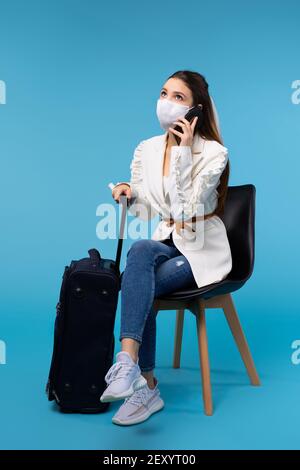 Junge Studentin trägt eine Schutzmaske und Gepäck sitzend Auf einem Stuhl spricht am Telefon kündigt Flugstreichungen an Aufgrund der Pandemie Stockfoto