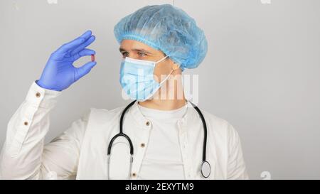 Ein gutaussehender junger Arzt untersucht eine neue Droge.EIN junger gutaussehender Arzt in einer medizinischen Maske hält eine rote Pille in seinen Händen und präsentiert eine neue Droge. Stockfoto