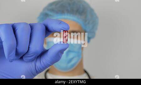 Ein gutaussehender junger Arzt untersucht eine neue Droge.EIN junger gutaussehender Arzt in einer medizinischen Maske hält eine rote Pille in seinen Händen und präsentiert eine neue Droge. Stockfoto